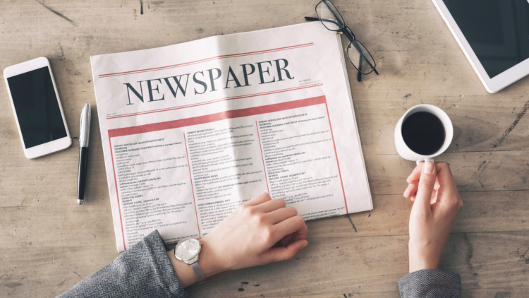 a person holds a cup of coffee and rests one hand on the front page of a newspaper